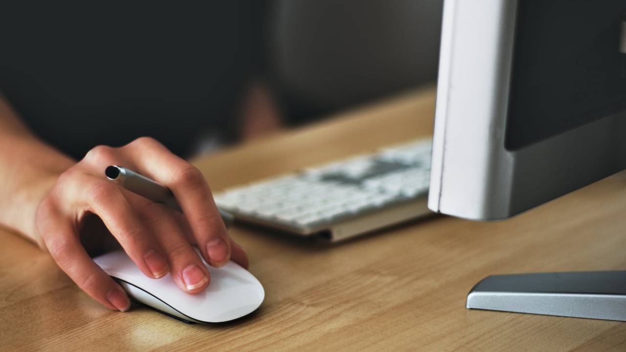 Imagem mostra uma mesa de trabalho de madeira. Sobre ela, uma pessoa mexe no mouse do computador e, na mesma mão, segura uma caneta.