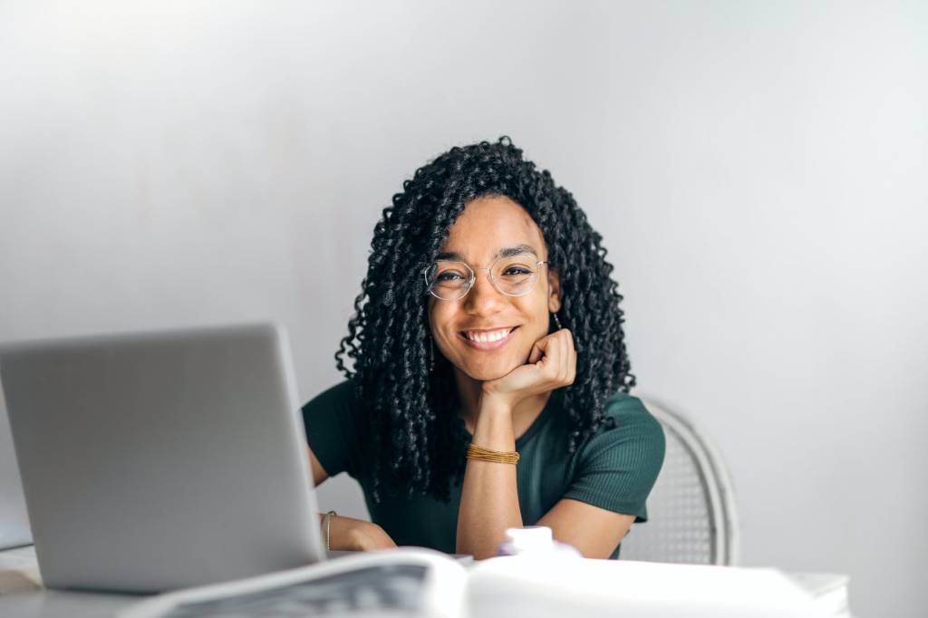 Jovem negra, com os cabelos cacheados, sorri para a imagem enquanto utiliza um computador