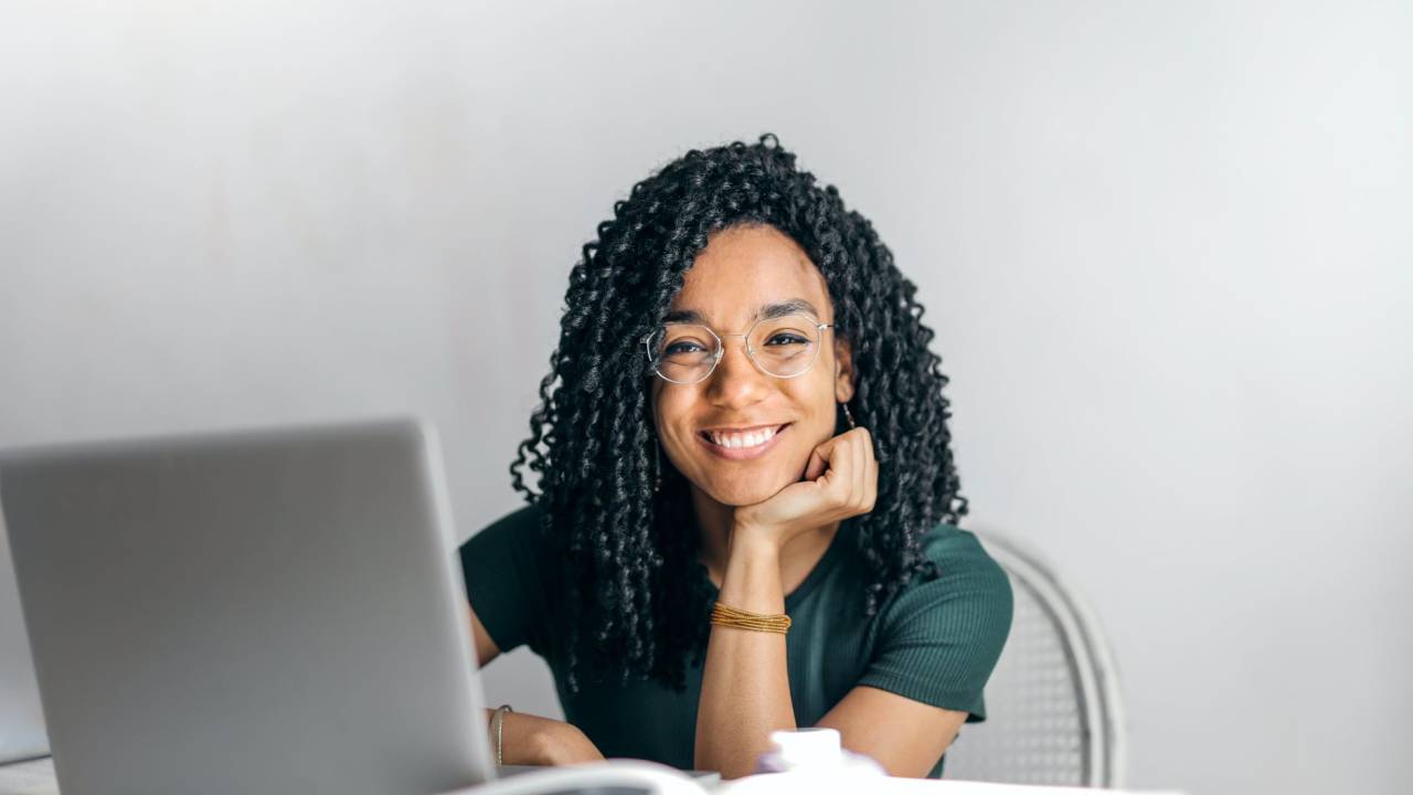 Jovem negra, com os cabelos cacheados, sorri para a imagem enquanto utiliza um computador