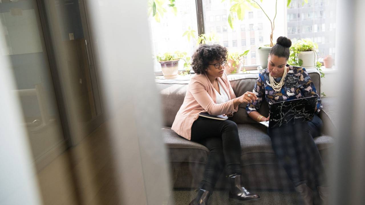 Duas mulheres negras estão sentadas em um sofá enquanto olham para a tela de um notebook que está no colo de uma delas.