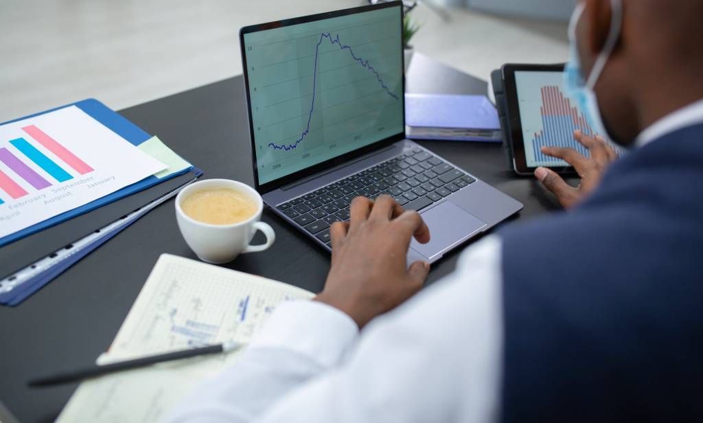 Imagem mostra um homem mexendo em um notebook e em um tablet ao mesmo tempo. Na tela de ambos há um gráfico. Sobre a mesa, além dos dois aparelhos, tem uma xícara de café e um caderno.