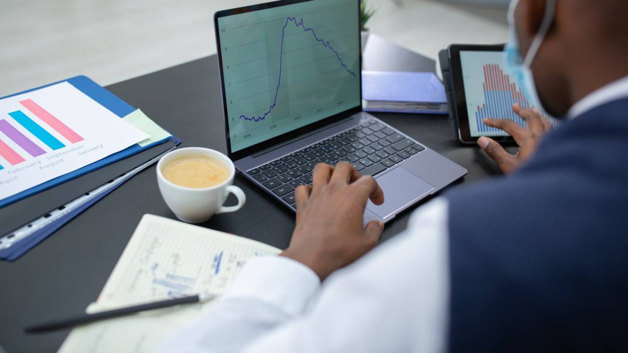 Imagem mostra um homem mexendo em um notebook e em um tablet ao mesmo tempo. Na tela de ambos há um gráfico. Sobre a mesa, além dos dois aparelhos, tem uma xícara de café e um caderno.