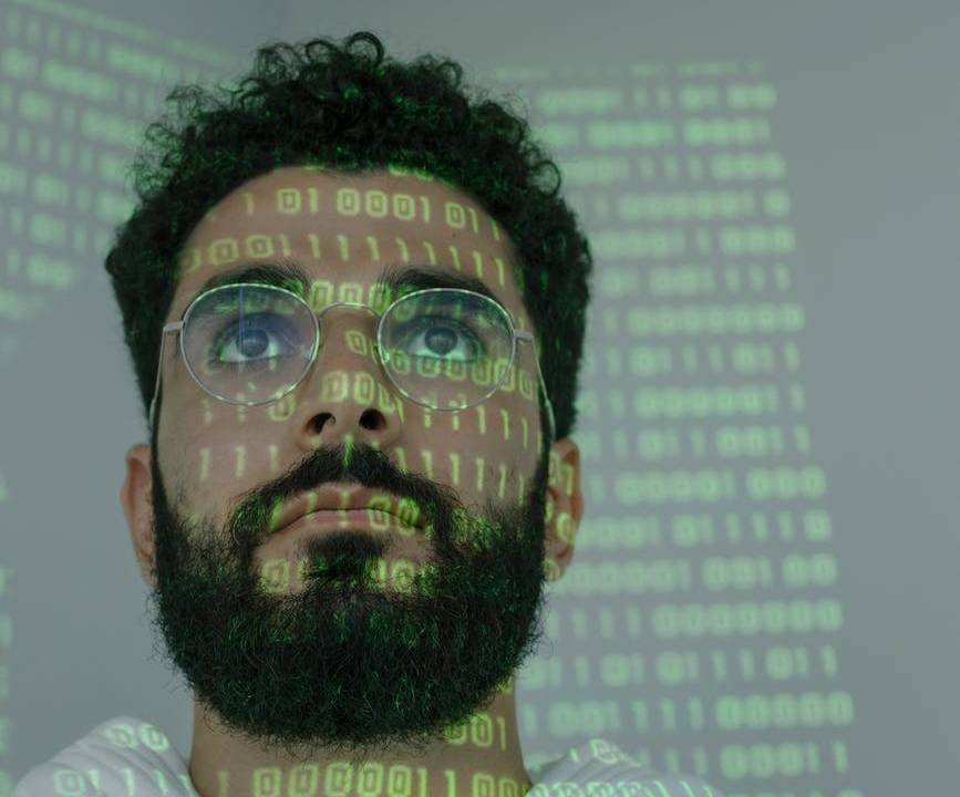 Imagem mostra o rosto de um homem olhando para uma tela. Em seu rosto, estão refletidos dados que estão na tela.