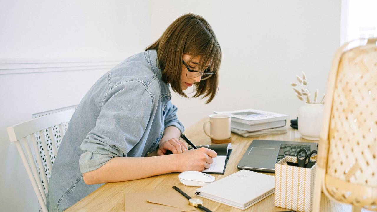 Mulher se debruça sobre uma mesa enquanto faz anotações em um caderno. Na mesa, há também um computador e demais itens de escritório.