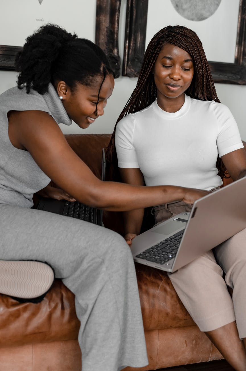Duas mulheres negras estão sentadas em um sofá, enquanto sorriem com o que veem na tela de um notebook