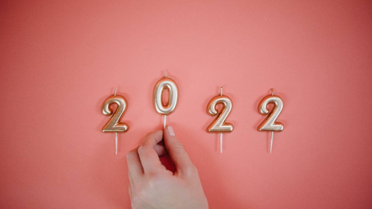 A imagem mostra um fundo cor de rosa e, por cima, quatro velas douradas que formam o ano de 2022. Uma mão aparece segurando o número zero