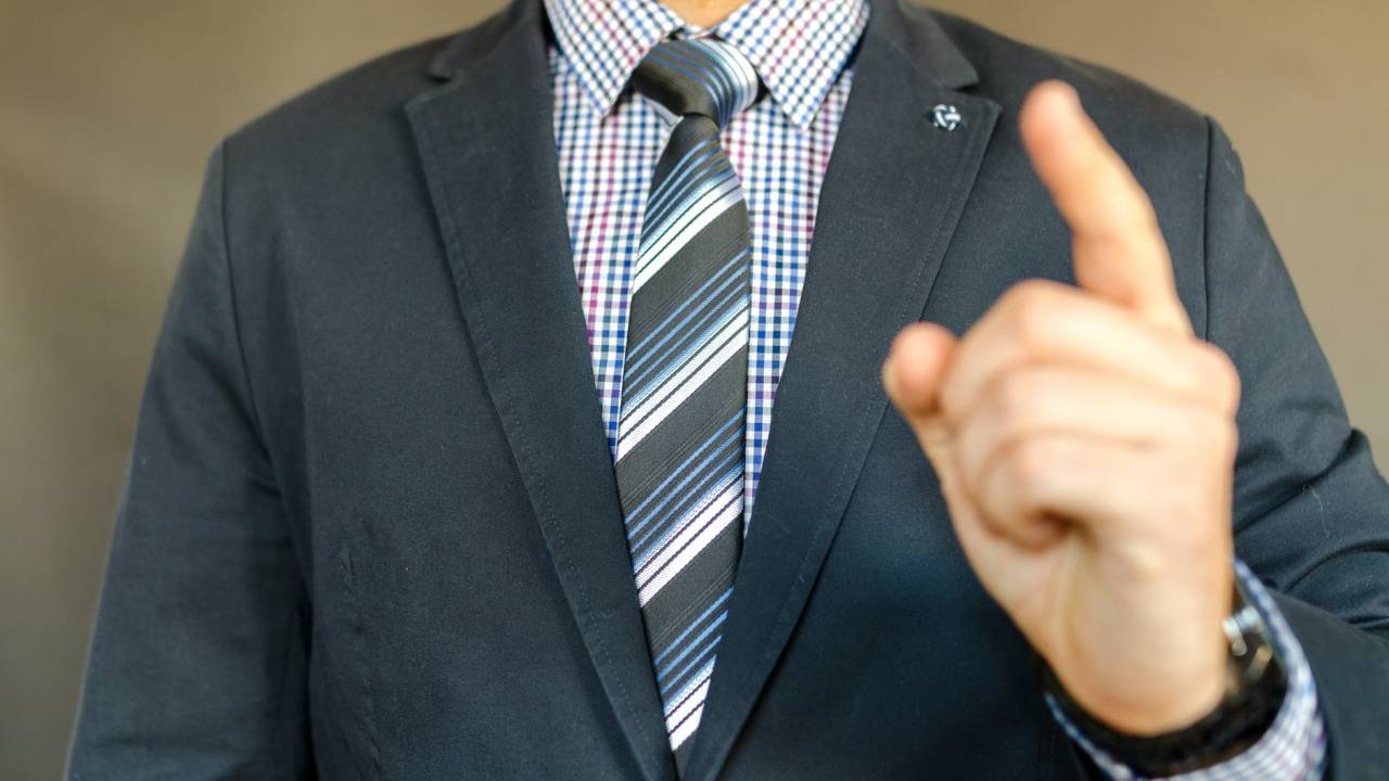 Homem de terno e gravata aponta o dedo em direção à câmera