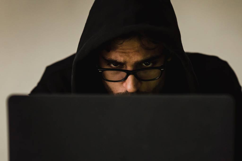 Imagem mostra um homem vestido com um moletom de capuz preto, de óculos, olhando para um computador. Apenas metade do rosto dele aparece na imagem.