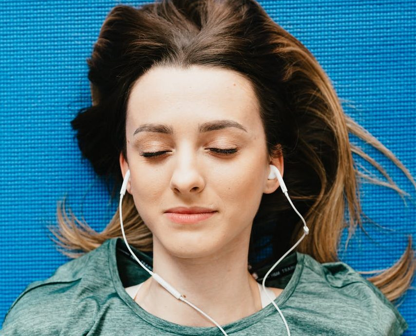 Imagem mostra uma mulher com fones de ouvido, deitada no chão sobre um tatame azul, de fones de ouvido e olhos fechados