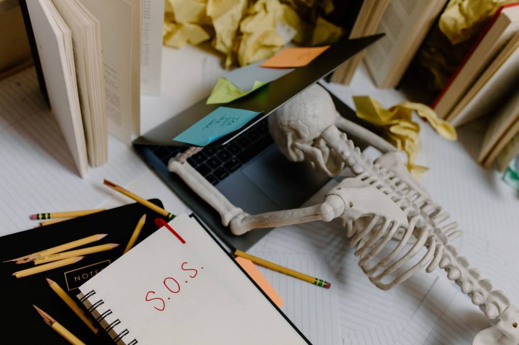 Imagem mostra um esqueleto apoiado sobre um notebook meio aberto. Ao lado, na mesa, tem canetas espalhadas e um caderno onde está escrito "S.O.S"