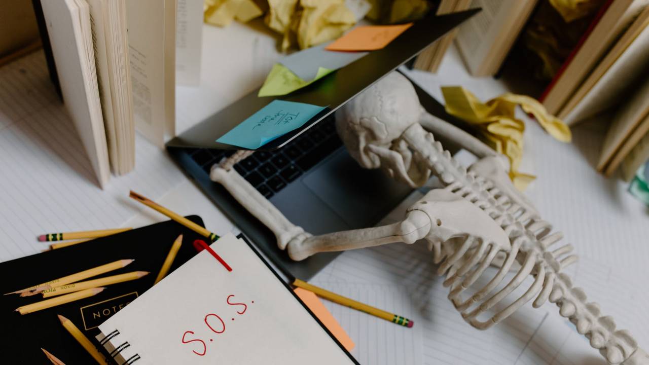 Imagem mostra um esqueleto apoiado sobre um notebook meio aberto. Ao lado, na mesa, tem canetas espalhadas e um caderno onde está escrito "S.O.S"