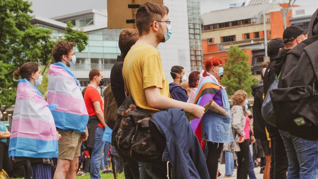 Imagem mostra várias pessoas reunidas ao ar livre. Elas usam máscara e algumas estão com bandeiras trans nos ombros