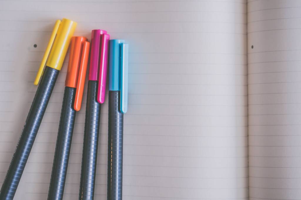 Imagem mostra uma folha de papel em branco e, sobre ela, quatro canetas: uma amarela, uma laranja, uma rosa e uma azul