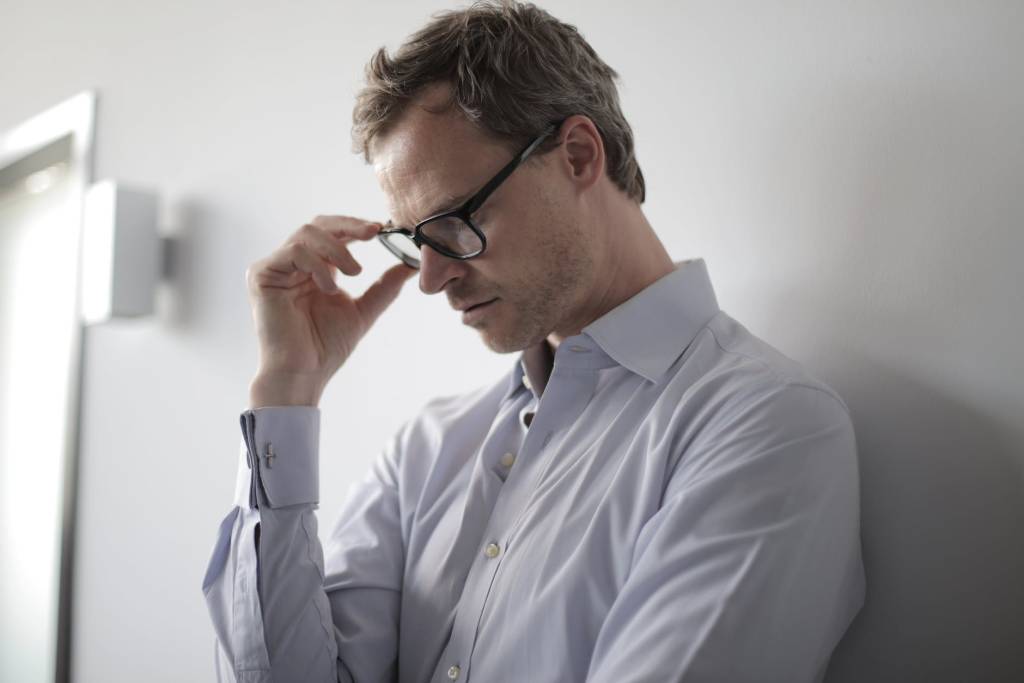 Imagem mostra um homem vestido com uma camisa branca encostado em uma parede. Ele segura os óculos e, de olhos fechados, tem um semblante preocupado