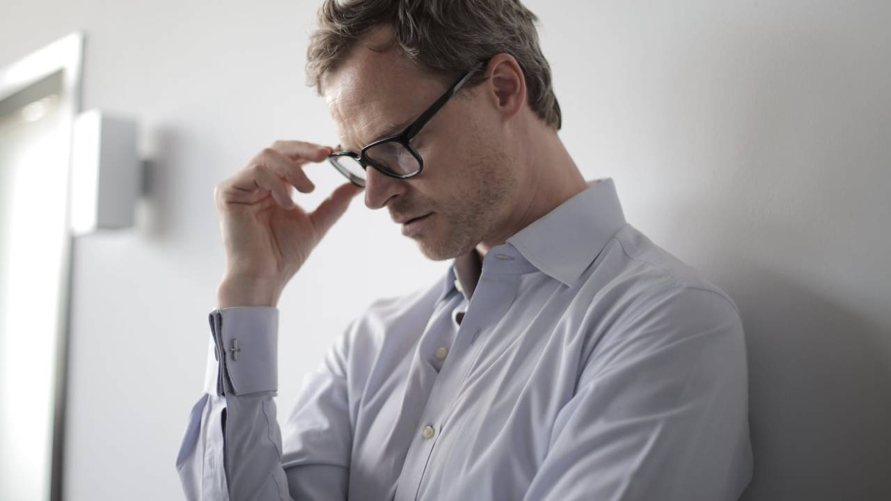 Imagem mostra um homem vestido com uma camisa branca encostado em uma parede. Ele segura os óculos e, de olhos fechados, tem um semblante preocupado