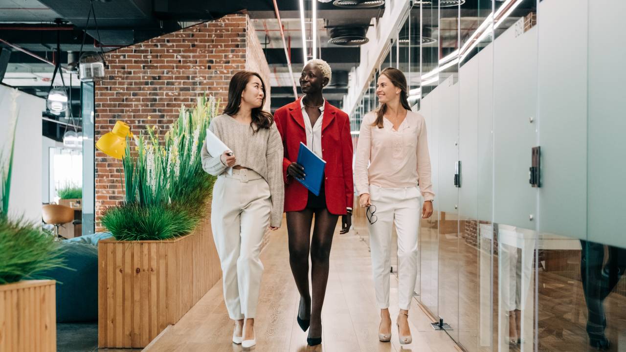 Três mulheres caminham no corredor de um grande escritório