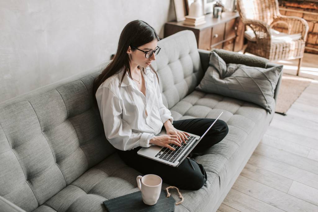Mulher está sentada sobre um sofá cinza, digitando no teclado de um notebook. Ao lado dela há uma xícara de chá