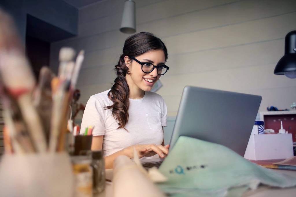 Uma mulher sorri enquanto olha para o computador. Suas mãos estão sobre o teclado e, sobre a mesa que está o notebook, há alguns materiais de escritório