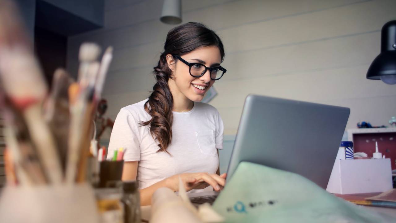 Uma mulher sorri enquanto olha para o computador. Suas mãos estão sobre o teclado e, sobre a mesa que está o notebook, há alguns materiais de escritório