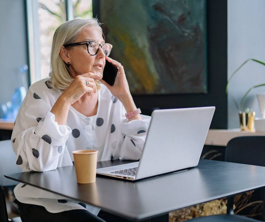 Uma mulher de cabelos brancos e óculos, vestida com uma camisa branca de bolas pretas, fala ao telefone enquanto trabalha