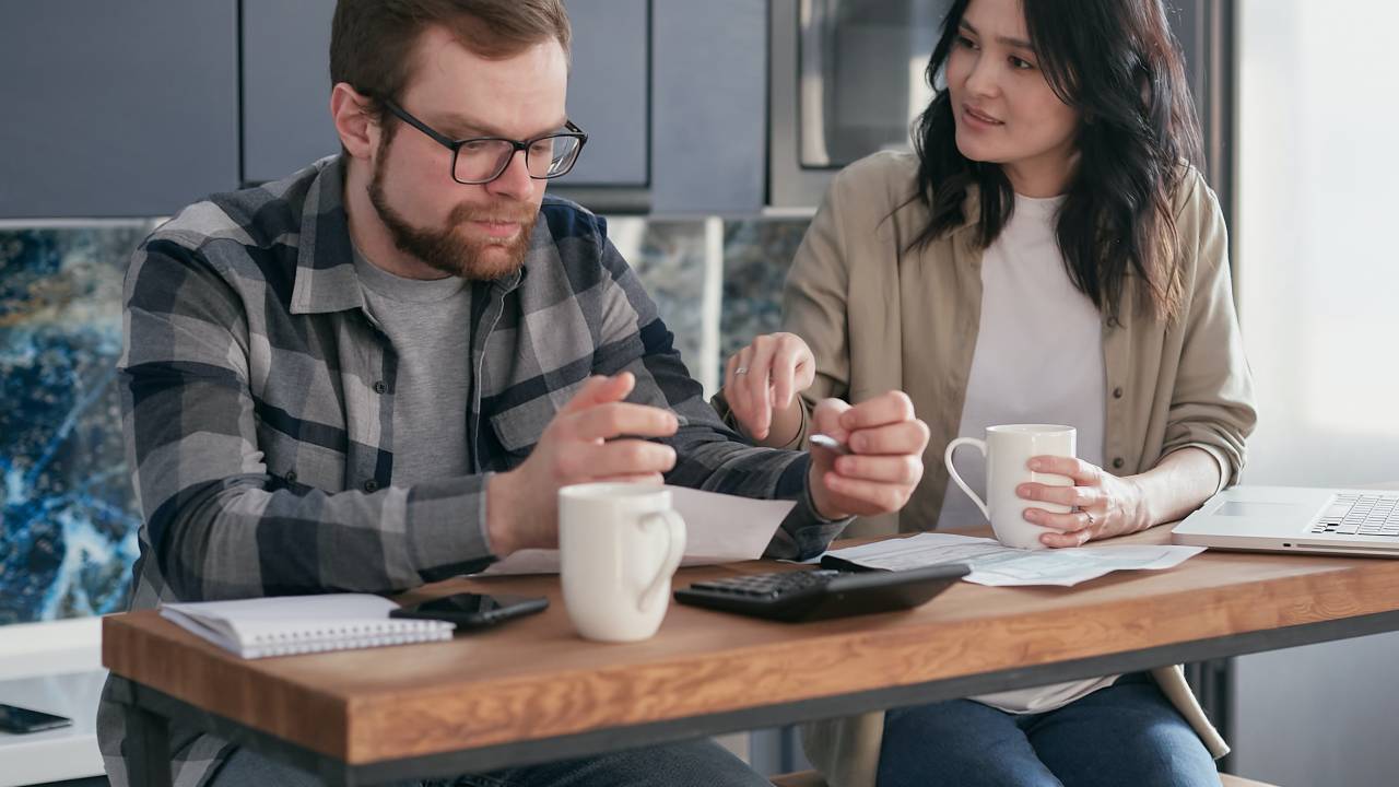 Um homem e uma mulher estão sentados à mesa, que tem duas xícaras, uma calculadora e alguns papéis espalhados. Os dois parecem preocupados
