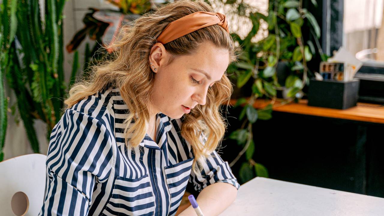Uma mulher está sentada enquanto olha para um caderno, concentrada, e segura um lápis