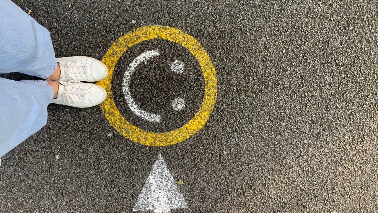 Imagem mostra parte das pernas e os pés de uma mulher com jeans e tênis branco próxima a um emoji sorrindo desenhando no asfalto