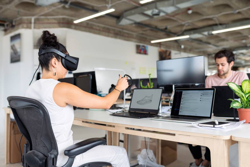 Uma mulher está sentada em uma cadeira em um ambiente corporativo, em frente a um computador, enquanto veste óculos de realidade virtual