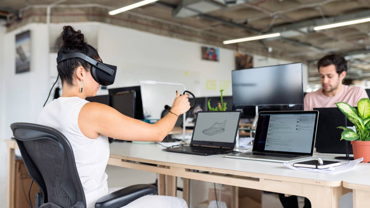 Uma mulher está sentada em uma cadeira em um ambiente corporativo, em frente a um computador, enquanto veste óculos de realidade virtual