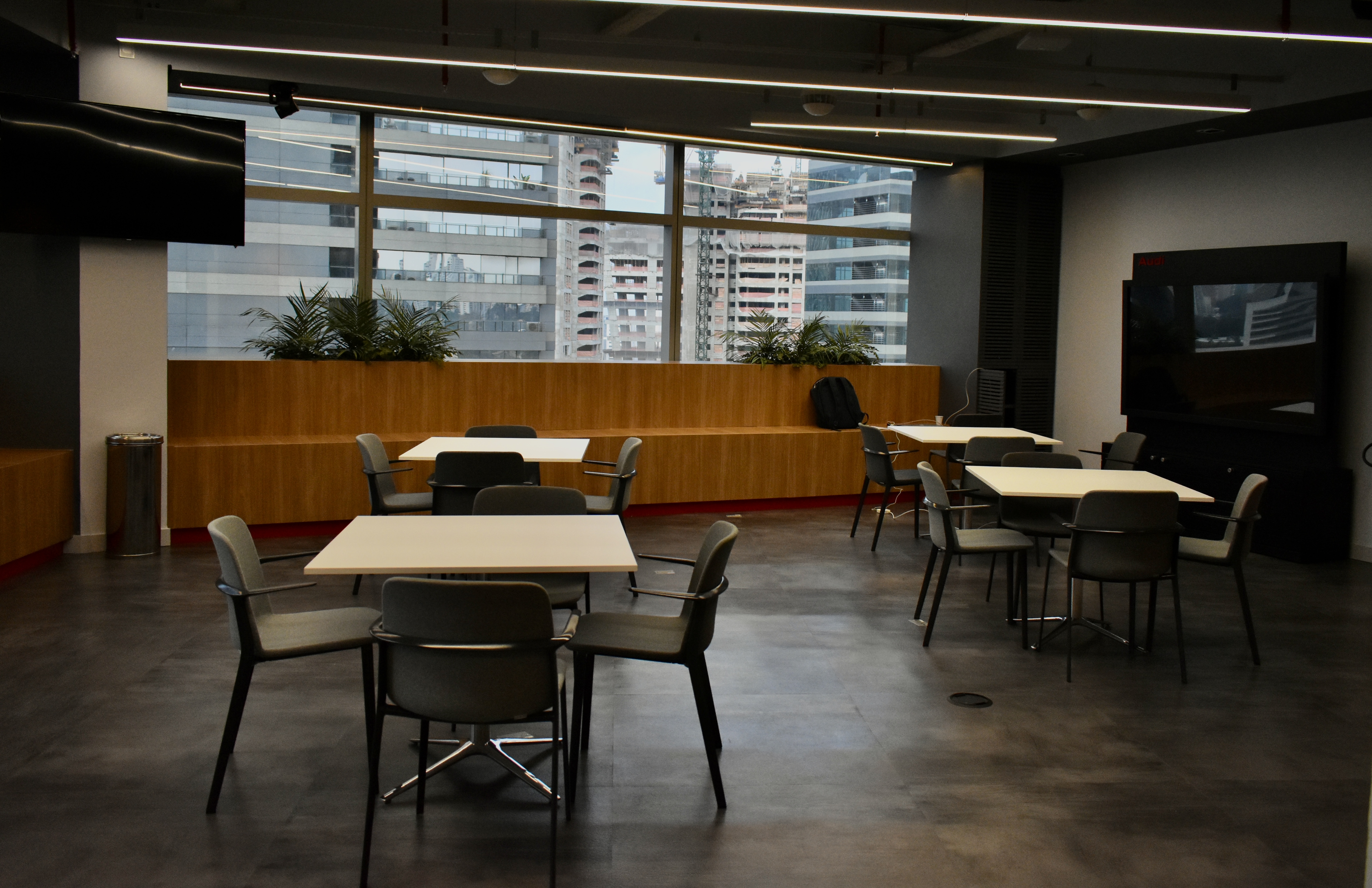 Imagem mostra a copa do novo escritório da Audi. O ambiente tem o piso de madeira, mesas de quatro lugares e, ao fundo, uma janela cuja vista são prédios de São Paulo