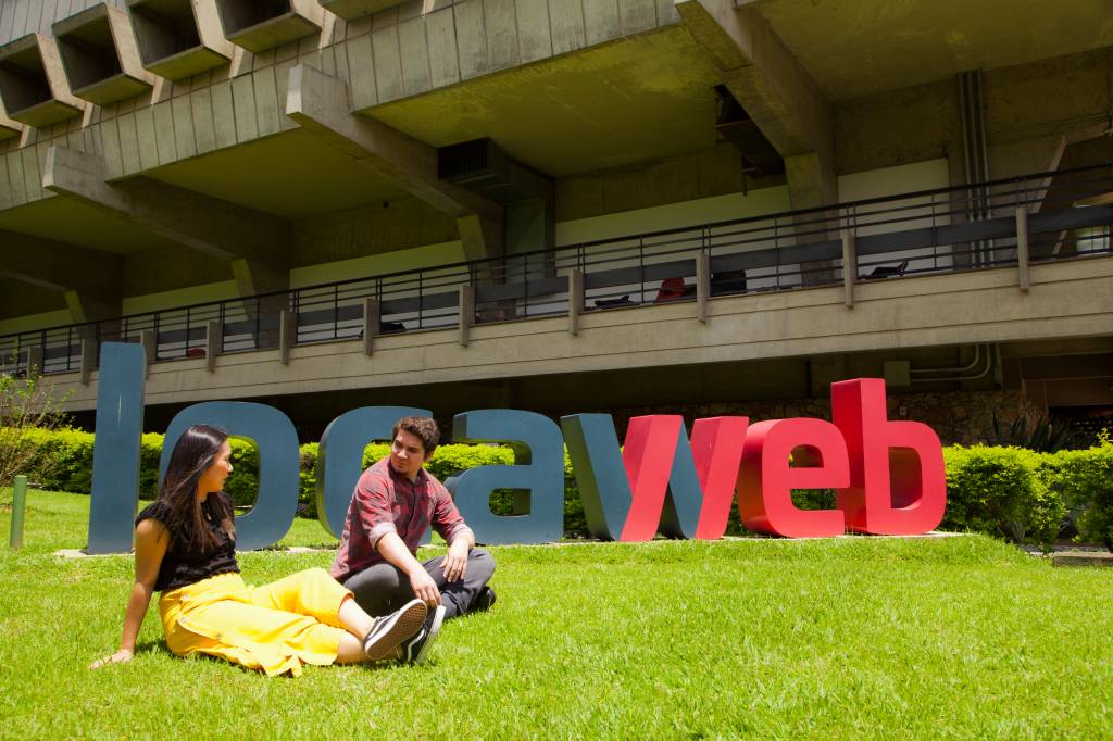 Um homem e uma mulher estão sentados em um gramado. Logo atrás deles há um letreiro onde está escrito Locaweb em azul e vermelho