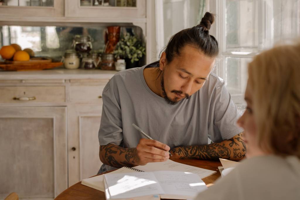Um homem vestido com uma camiseta cinza, com tatuagens no braço e um coque no cabelo está concentrado enquanto faz anotações em um caderno