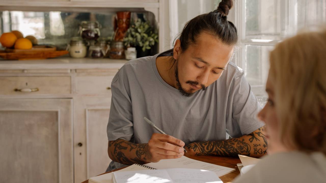 Um homem vestido com uma camiseta cinza, com tatuagens no braço e um coque no cabelo está concentrado enquanto faz anotações em um caderno