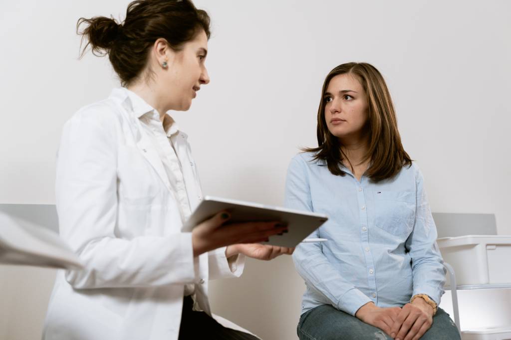 Uma mulher está sentada em uma maca enquanto conversa com uma médica. A médica, vestida de jaleco, está com uma prancheta na mão