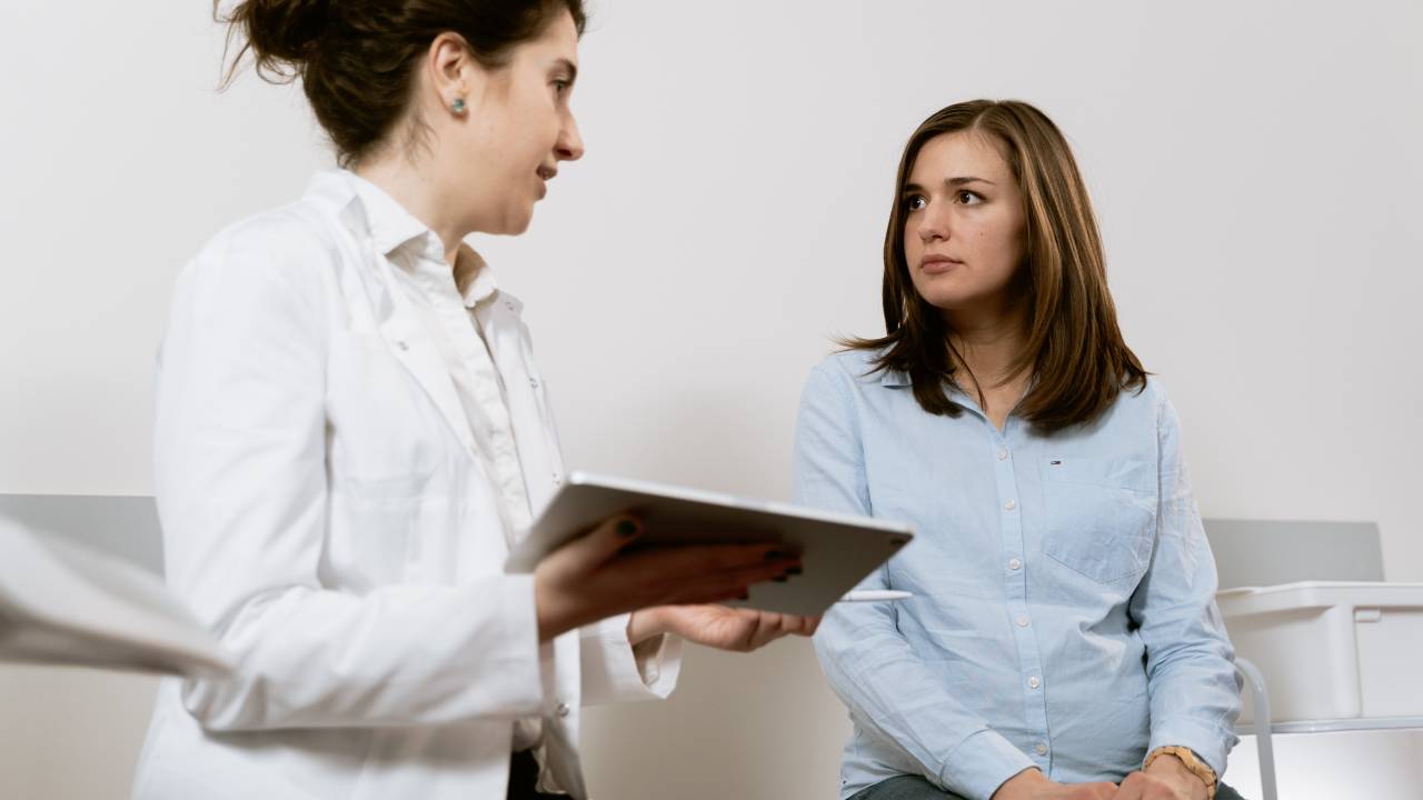 Uma mulher está sentada em uma maca enquanto conversa com uma médica. A médica, vestida de jaleco, está com uma prancheta na mão