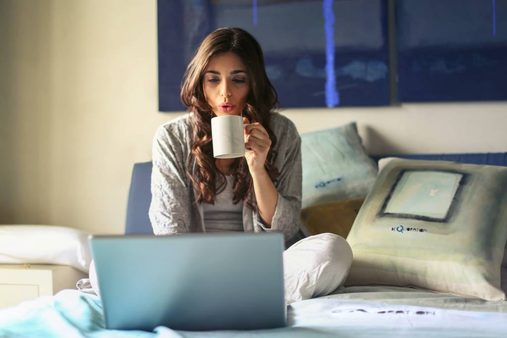 Uma mulher de cabelo longo e castanho está sentada sobre uma cama enquanto toma algo em uma xícara e olha para a tela de um notebook
