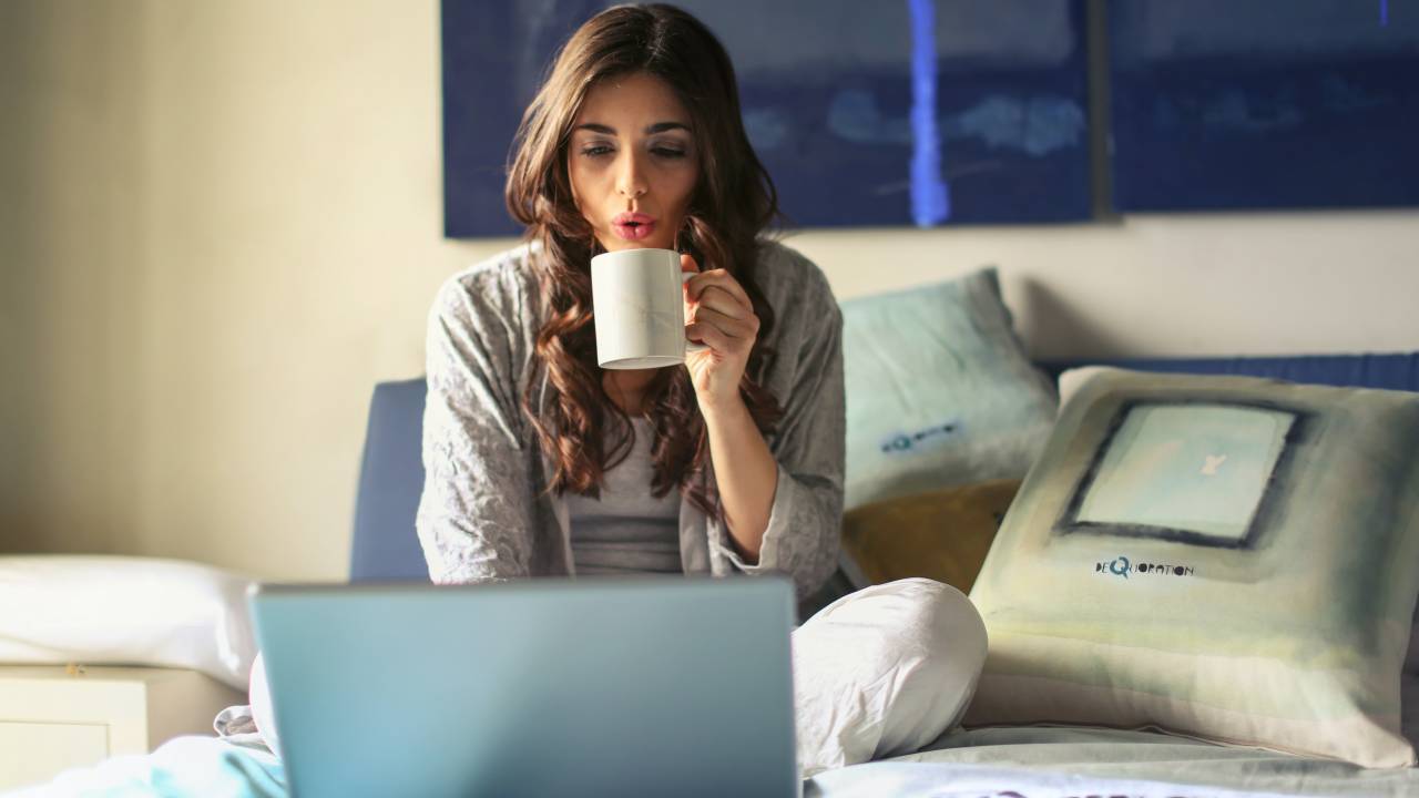 Uma mulher de cabelo longo e castanho está sentada sobre uma cama enquanto toma algo em uma xícara e olha para a tela de um notebook