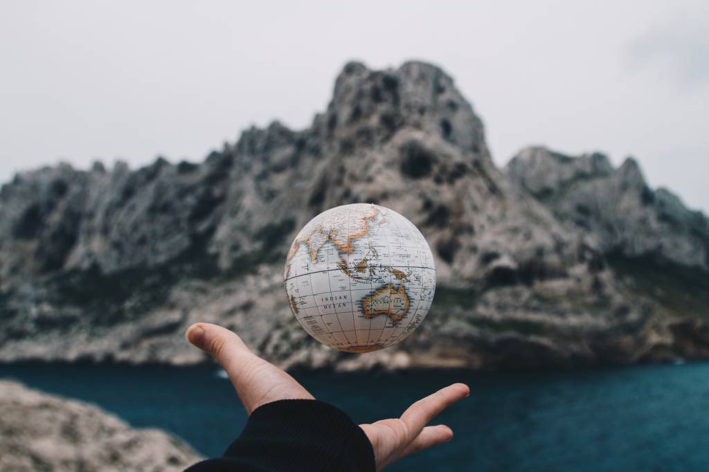 Em uma paisagem montanhosa e fria, uma mão segura um globo terrestre. A foto é feita no momento em que o globo é lançado para cima pela mão.