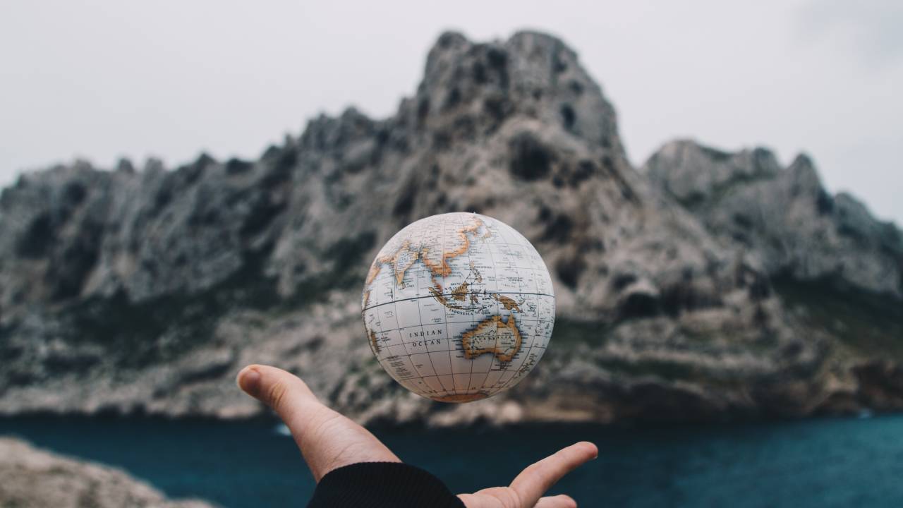 Em uma paisagem montanhosa e fria, uma mão segura um globo terrestre. A foto é feita no momento em que o globo é lançado para cima pela mão.