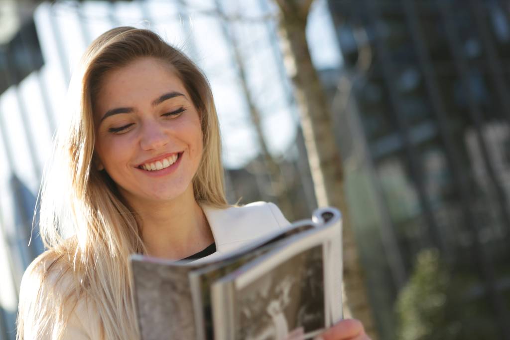 Uma mulher está em um ambiente ao ar livre lendo um livro enquanto sorri