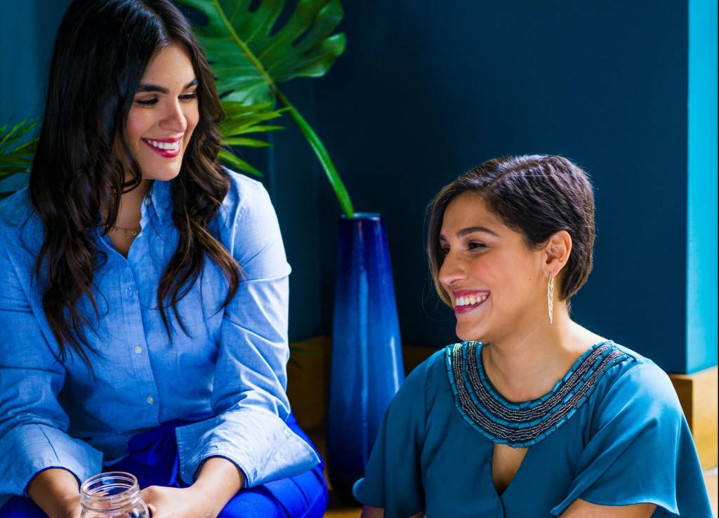 Duas mulheres conversam entre si. As duas estão sentadas e vestem tons entre azul e verde. Atrás delas, há um vaso com uma planta. Elas estão em um ambiente fechado.