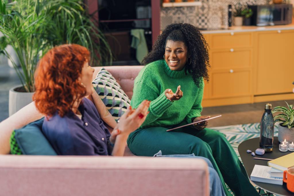 Duas mulheres, uma negra e uma ruiva, estão sentadas em um sofá conversando e sorrindo