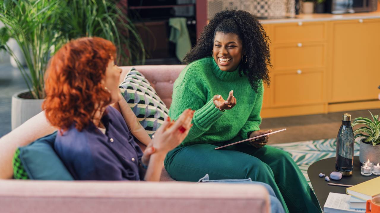 Duas mulheres, uma negra e uma ruiva, estão sentadas em um sofá conversando e sorrindo