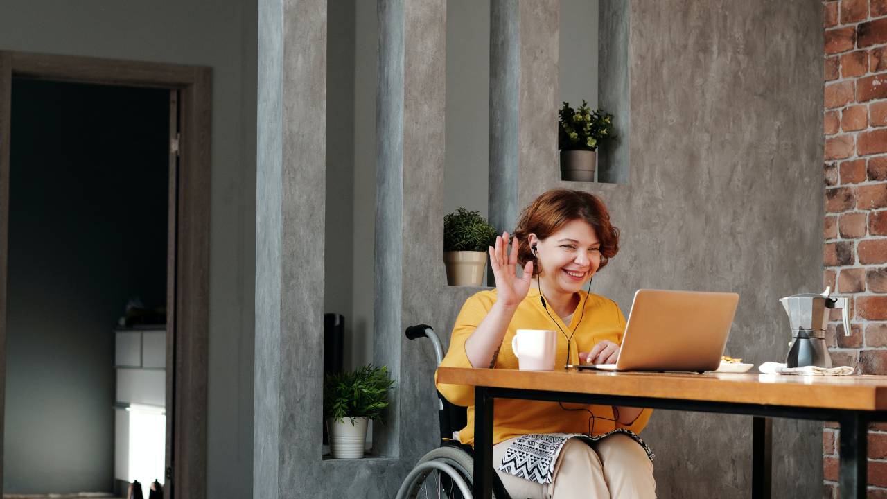 Uma mulher está sentada sobre uma cadeira de rodas. Ela veste uma blusa amarela e uma calça branca. Ela está em frente a um computador e interage com alguém por meio dele.