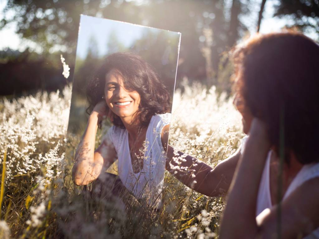 Mulher sentada ao gramado segura um espelho e vê sua imagem sorridente refletida nele