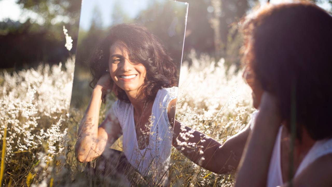 Mulher sentada ao gramado segura um espelho e vê sua imagem sorridente refletida nele