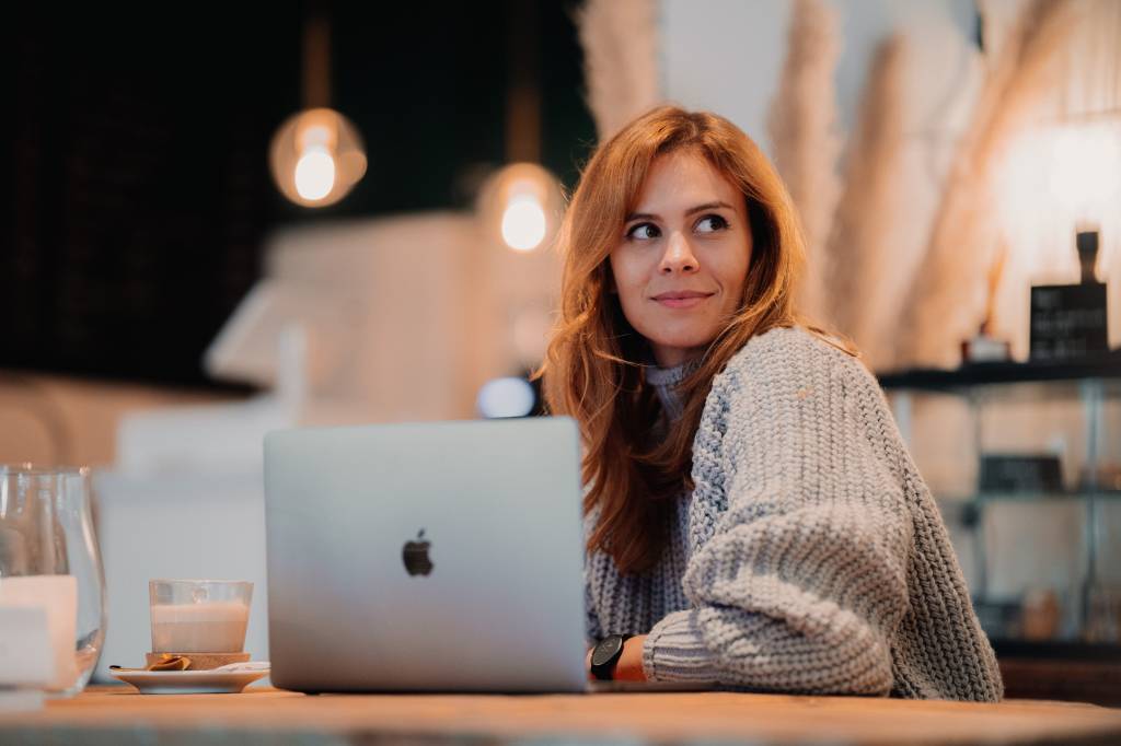 Mulher com cabelo ruivo comprido e blusa de lã sorri em frente a um notebook da Apple