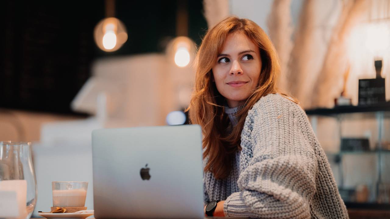 Mulher com cabelo ruivo comprido e blusa de lã sorri em frente a um notebook da Apple