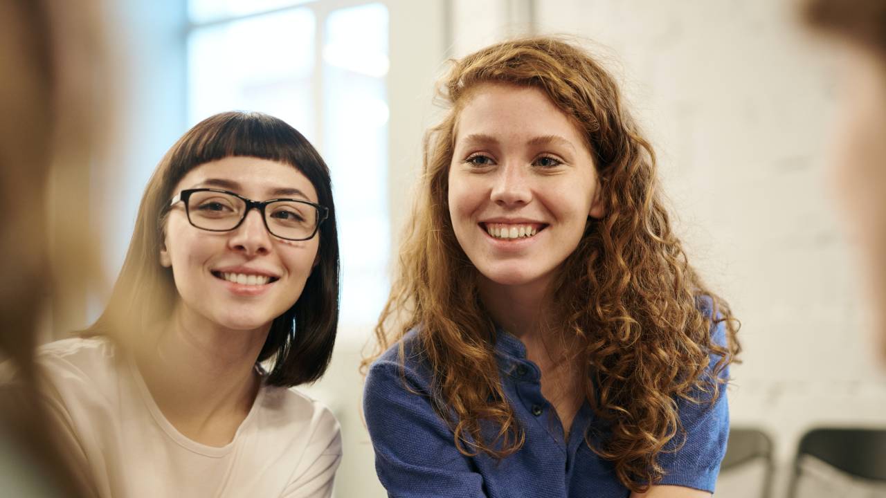 Duas mulheres, uma vestindo uma camiseta branca e óculos, e a outra usando uma camisa azul, sorriem para a foto.