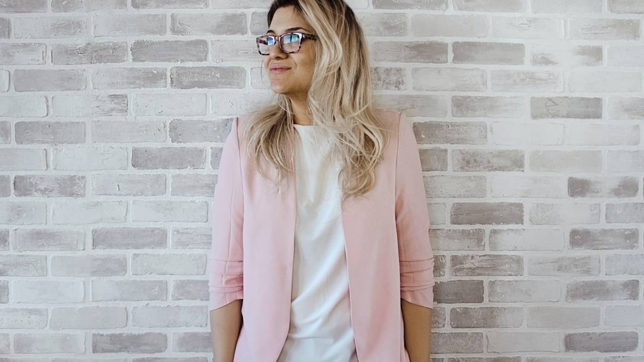 Mulher de blazer cor-de-rosa, camiseta branca e calça jeans, com óculos tartaruga, está em frente a uma parede de tijolinhos